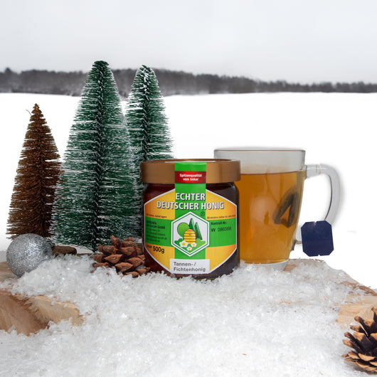 Ein Glas Honig auf einem Holzbrett. Im Hintergrund eine Schneelandschaft.